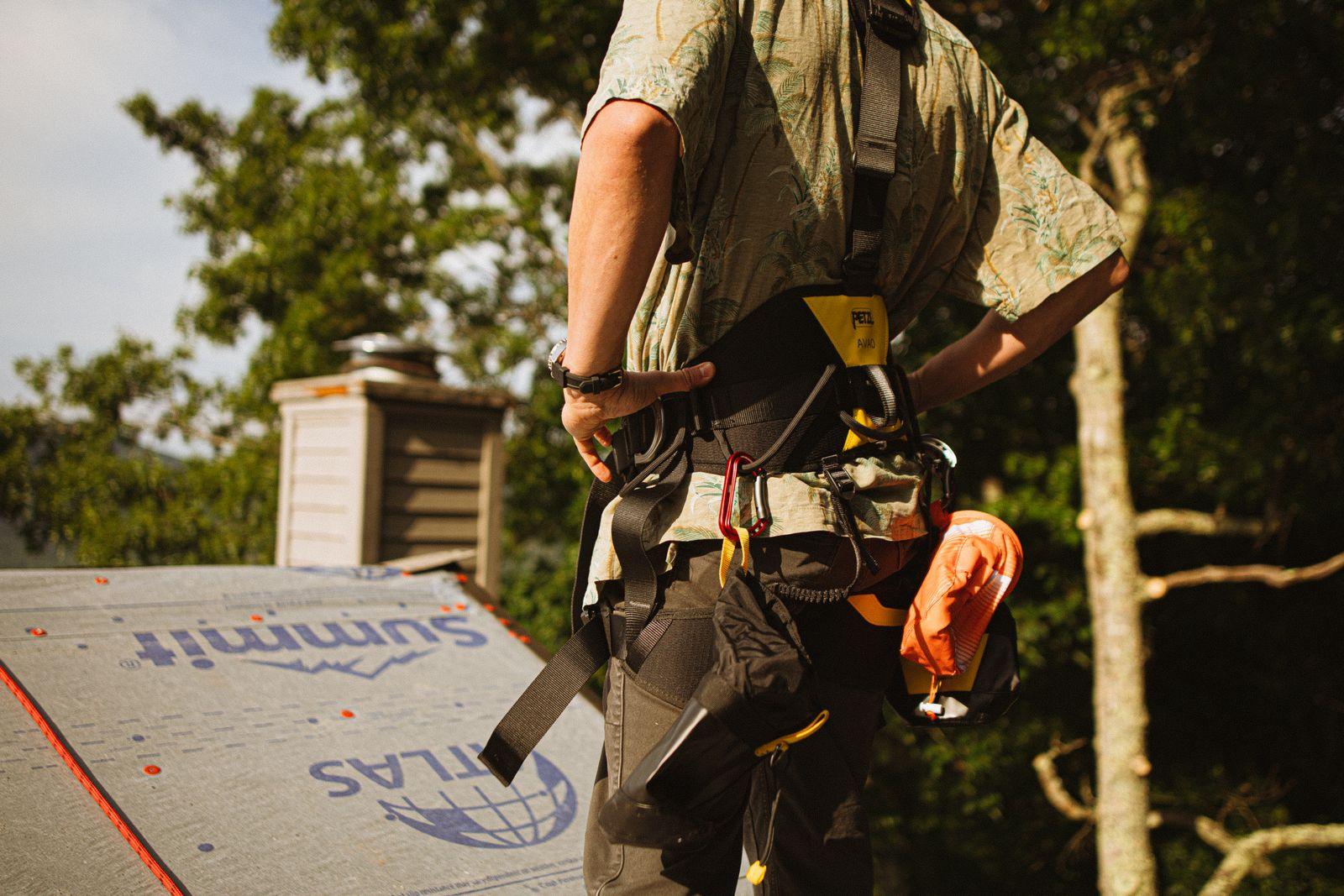 A roofer in a safety harness working on the shingles of a residential roof.