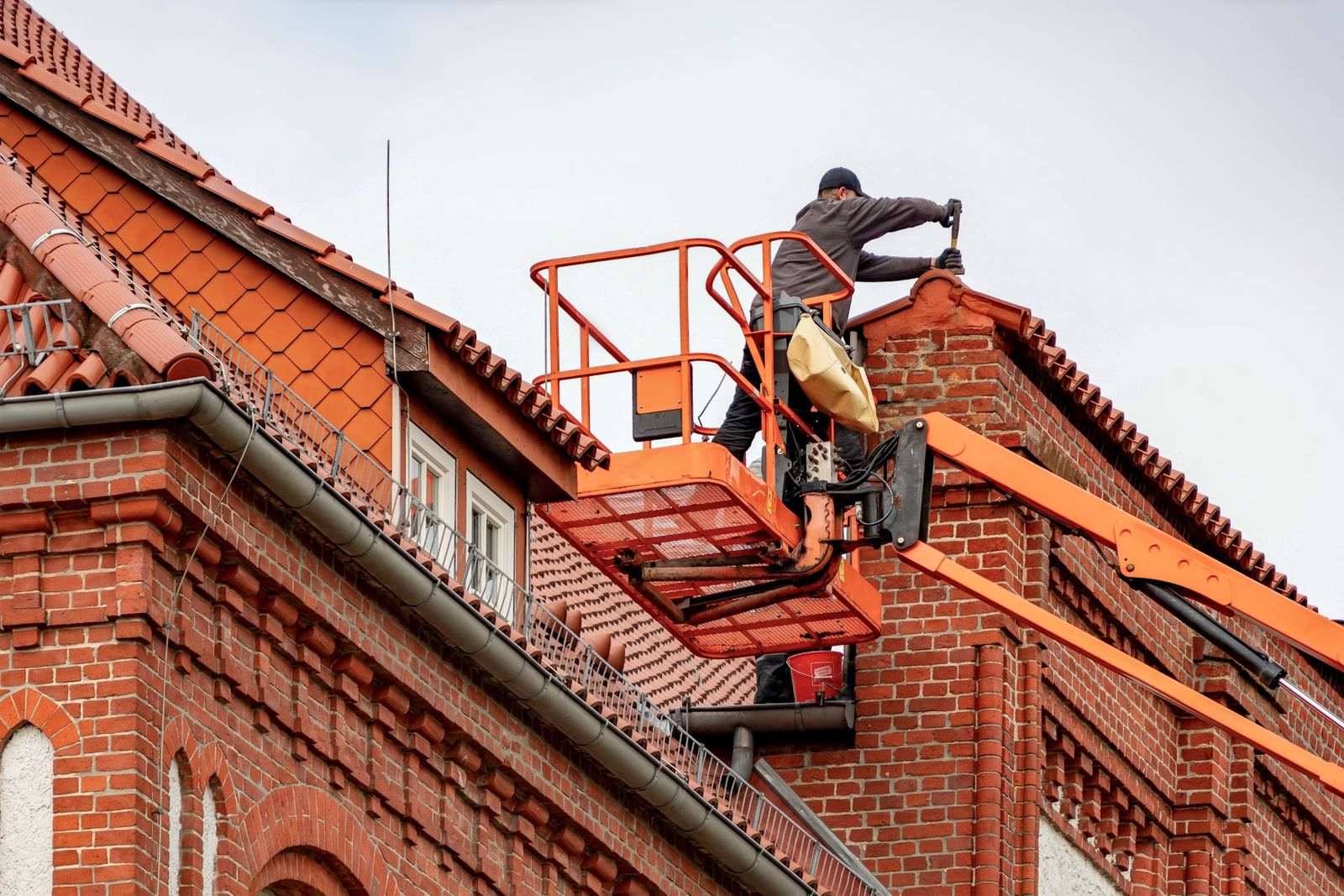 A worker on a bucket lift fitting a gutter section near the roof eave of a brick house
